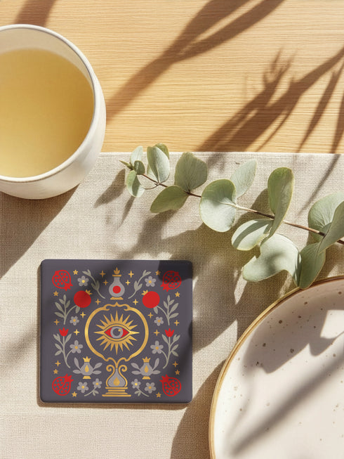 A square ceramic drink coaster with a smooth, matte charcoal grey finish. The surface features a symmetrical, intricate design in metallic gold, deep garnet red, and light grey. In the center is a golden All-Seeing Eye with a red iris, radiating light. The eye is framed by a golden circle and flanked by botanical elements, including red pomegranates and grey flowers with gold details. The design has a folk art, gothic botanical style.